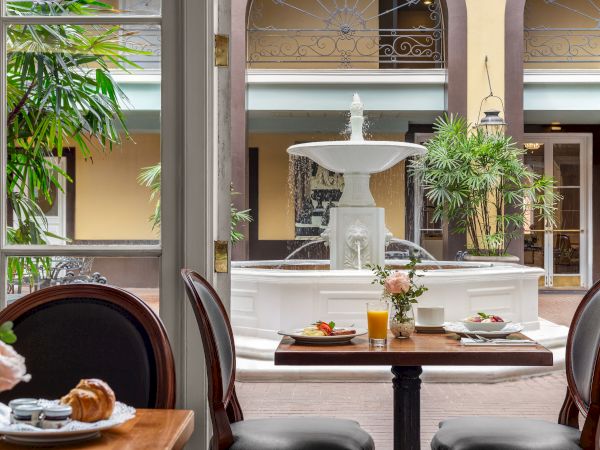 A charming courtyard with a central fountain, viewed from a dining area with tables set for breakfast, including juice and pastries.