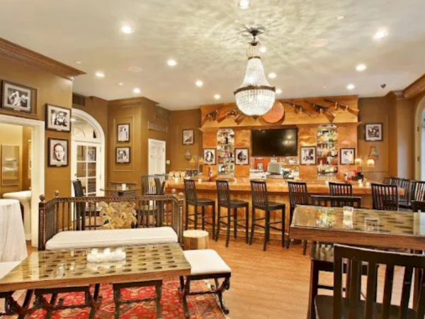 A cozy dining room or cafe with warm lighting, wooden tables and chairs, a chandelier, and framed photos on the tan walls.