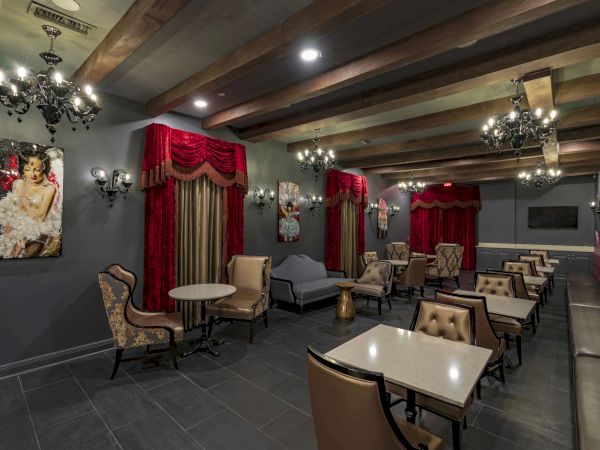 A stylish, dimly lit lounge with red drapes, chandeliers, and booth seating; tables and chairs arranged for socializing in a sophisticated setting.