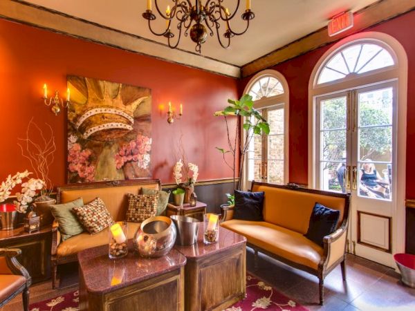 A cozy, red-walled lounge with leather sofa, coffee table, plants, petals, and warm chandelier lighting by arched windows, inviting and chic.