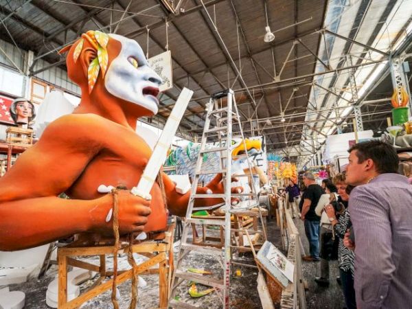A man wearing a large orange and white mask is sculpting a white torches or wand in a busy workshop with ladders, tools, and onlookers.