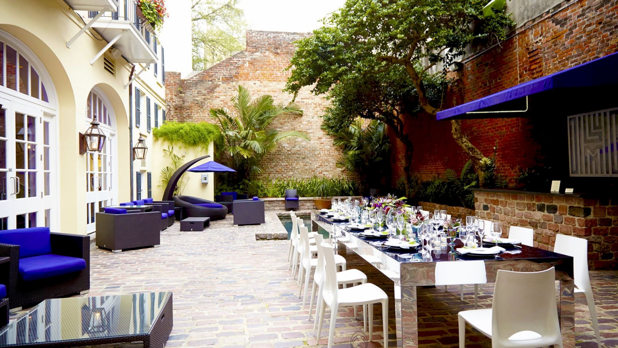An outdoor patio with a set dining table, modern furniture, plants, and trees against a brick wall backdrop.