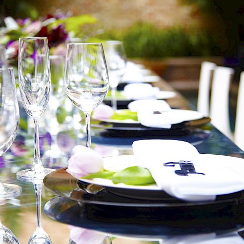 A long dining table is elegantly set with plates, wine glasses, and napkins, with floral decorations.