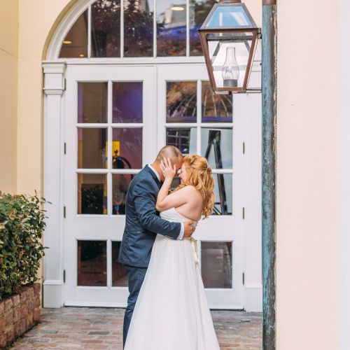 A couple in wedding attire embraces in front of a building with a large door and lantern, standing on a brick pathway.