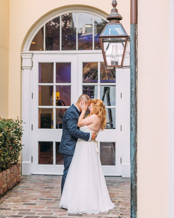 A couple in wedding attire embraces in front of a building with a large door and lantern, standing on a brick pathway.