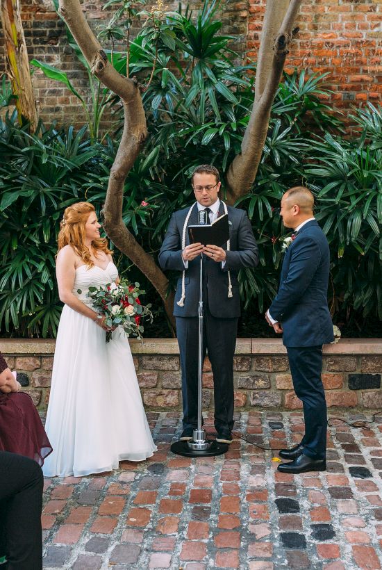A wedding ceremony in a garden setting with a couple, officiant, and guests seated on white chairs.