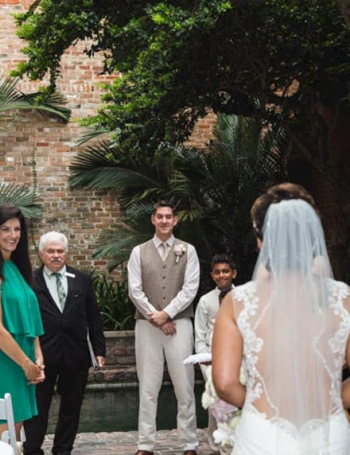 A wedding scene with a bride facing guests and a groom. Guests are dressed in various colors with greenery and a brick wall background.