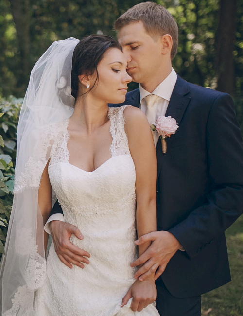 A bride and groom in wedding attire share a tender kiss in a garden, holding hands with a pink bouquet nearby.