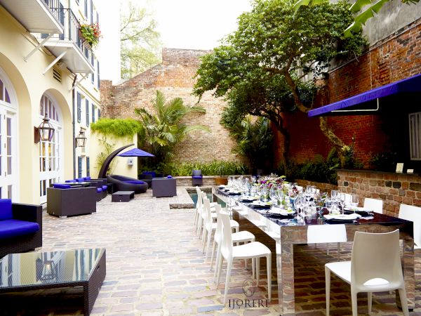 An outdoor courtyard event setup with a long table, white chairs, glassware, colorful flowers, and lush plants beside brick walls.