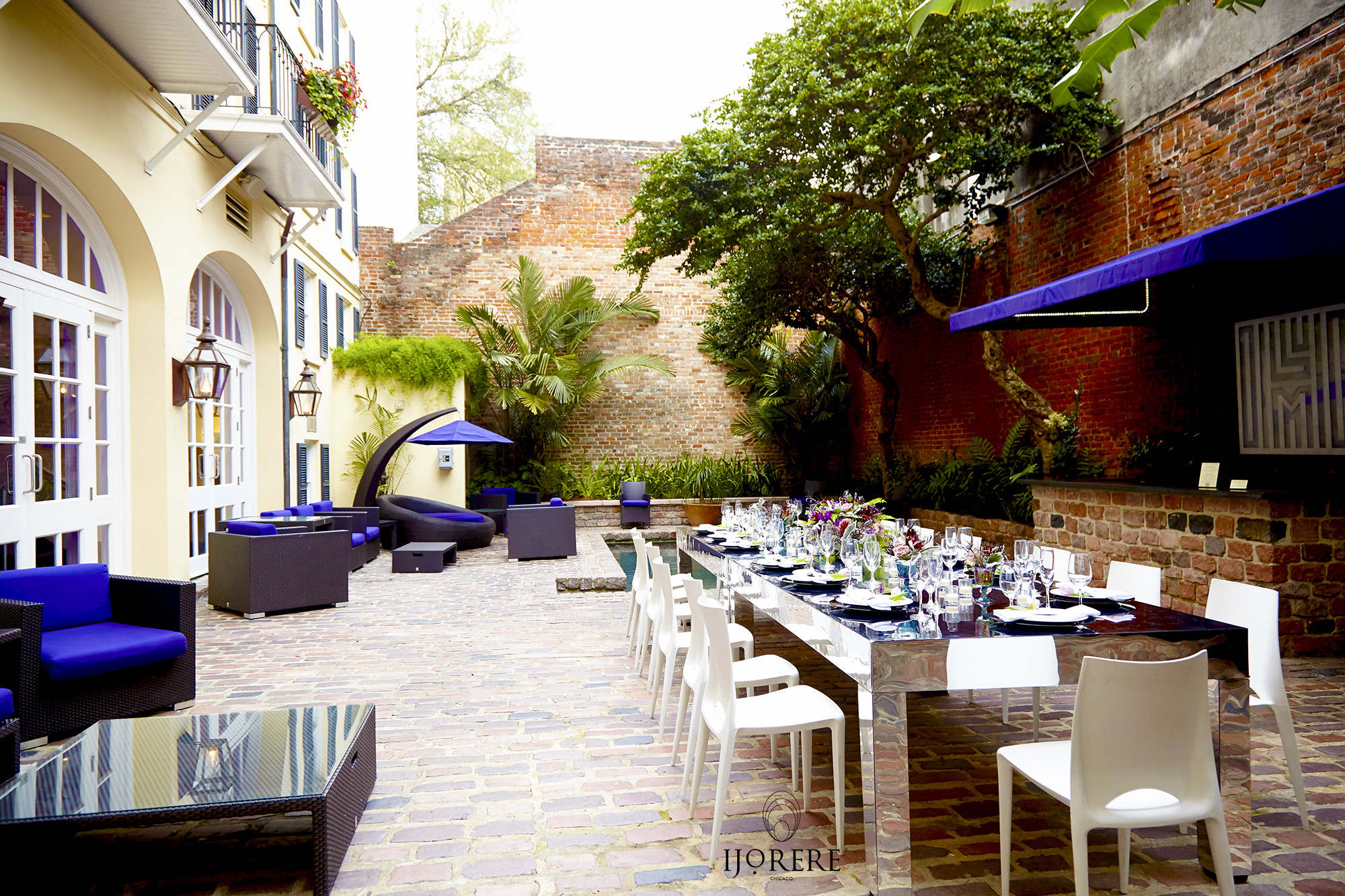A beautifully set outdoor dining area with a long table and white chairs, surrounded by brick walls and plants.