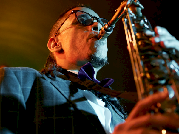 A saxophonist wearing a suit and bow tie, performing on stage with a microphone and warm concert lighting.