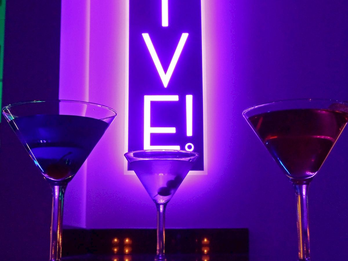 A neon vertical sign spells &ldquo;VIVE&rdquo; in purple light, flanked by three cocktail glasses on a bar counter.