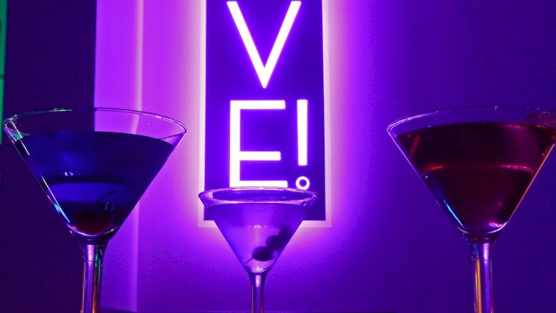Three martini glasses on a bar with a neon sign in the background.