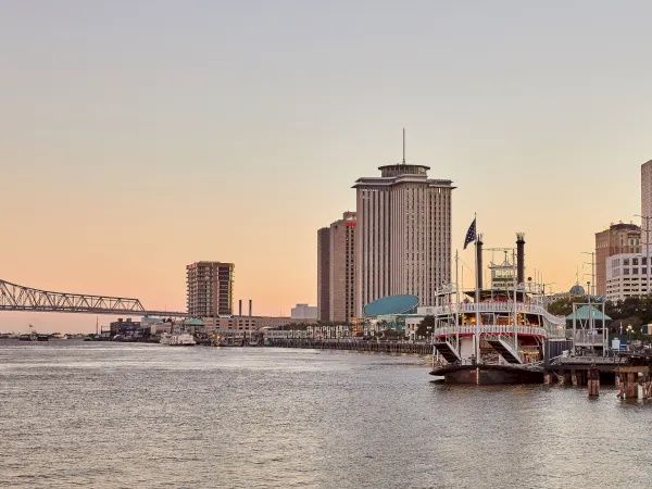 City skyline on a river at sunset, with a tall skyscraper and a docked boat along the waterfront.