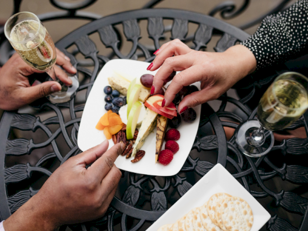 Two people are plating a fresh fruit and veggie lunch on a white plate over an outdoor wrought-iron table, toasting with drinks.