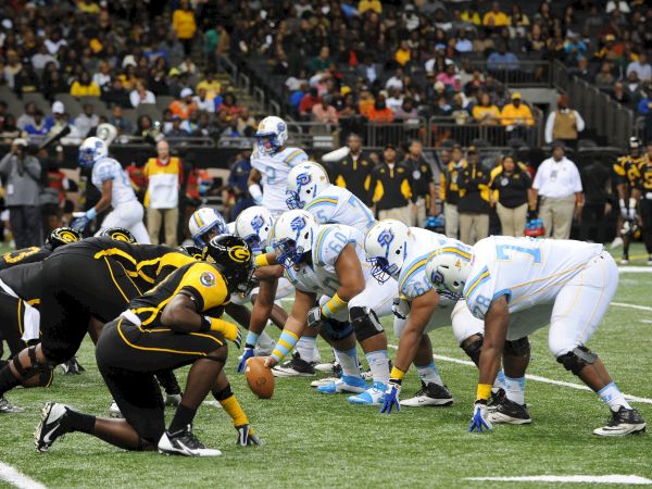 Players clash in a tense American-football scrum&mdash;white-and-blue offense meets black-and-yellow defense at the line.