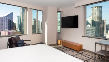 A modern hotel room with a bed, TV, and large windows offering a city skyline view, complete with a chair and table.