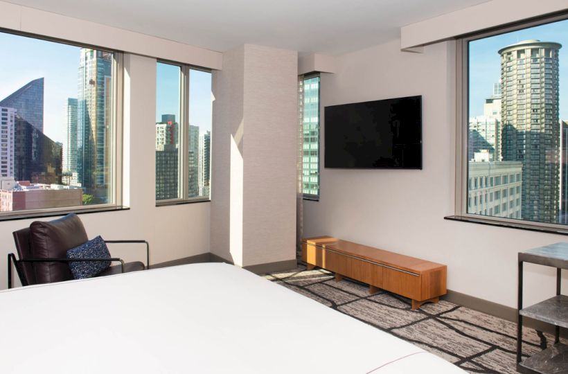 A modern hotel room with a bed, TV, and large windows offering a city skyline view, complete with a chair and table.