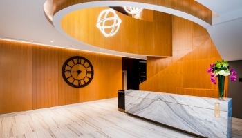 A modern lobby with a spiral wooden structure, clock, marble desk, and decorative lights and flowers.