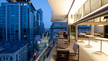 Rooftop terrace with seating overlooks a cityscape of modern buildings at dusk, featuring bright indoor lighting and a clear glass railing.