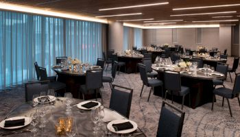 The image shows an elegantly set dining area with round tables, chairs, glassware, and flowers, under softly lit modern ceiling lights.