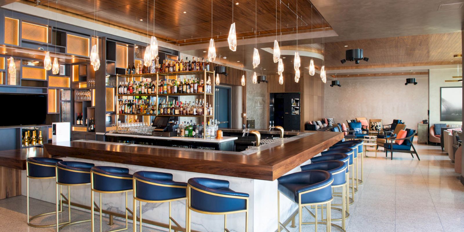 A modern bar with blue stools, elegant pendant lights, and a fully stocked liquor shelf in a stylish, spacious setting.