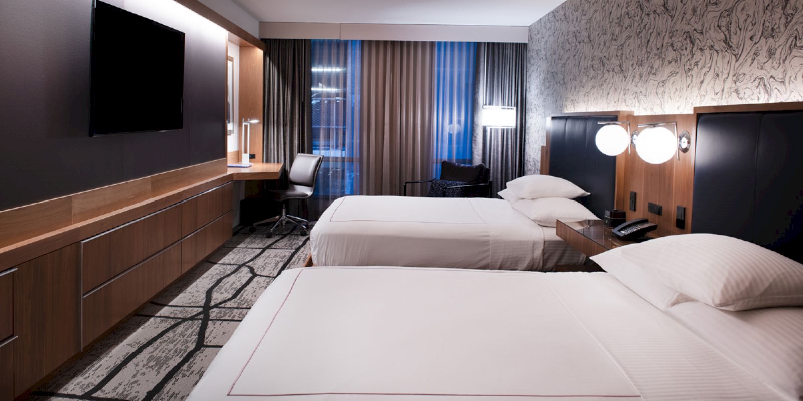 The image shows a hotel room with two beds, a desk with a chair, a TV on the wall, and modern decor and lighting.
