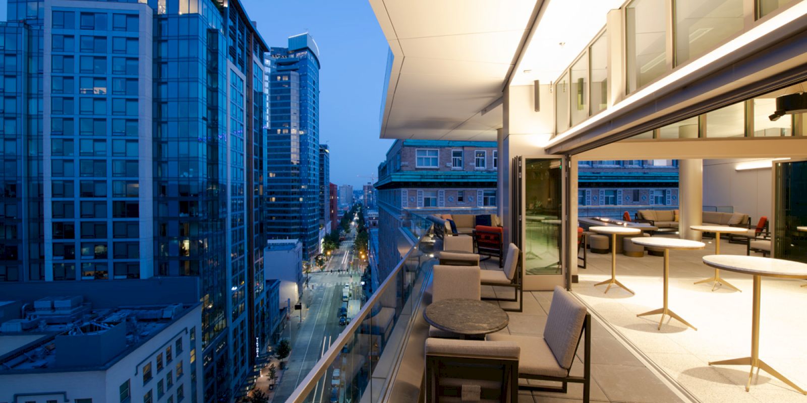 A modern rooftop terrace featuring seating, tables, and a view of a cityscape with tall buildings at dusk.