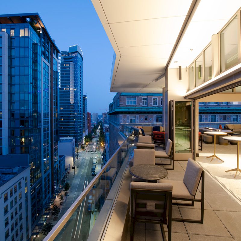 Rooftop patio with modern seating overlooks a vibrant cityscape at dusk, featuring high-rise buildings and illuminated streets.