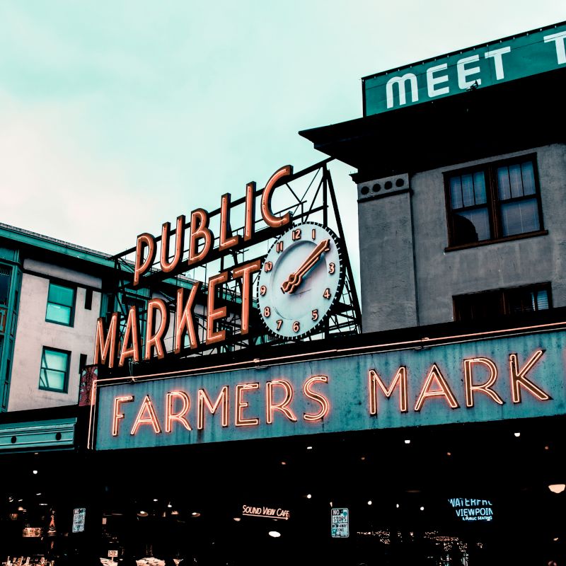 The image shows a public market with a large clock, neon signs for "Farmers Market," and buildings in the background, under a cloudy sky.