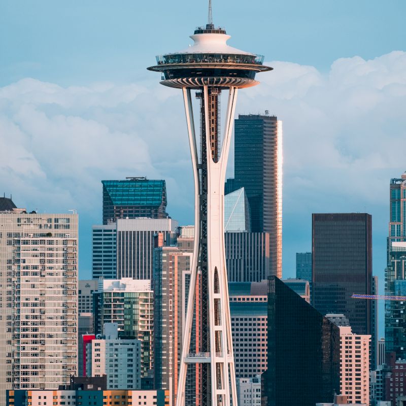 The image shows the Space Needle in Seattle with a backdrop of surrounding buildings under a partly cloudy blue sky.