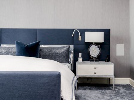 A modern bedroom interior with a blue headboard, white bedding, nightstand, lamp, decorative stone, and grey carpet.