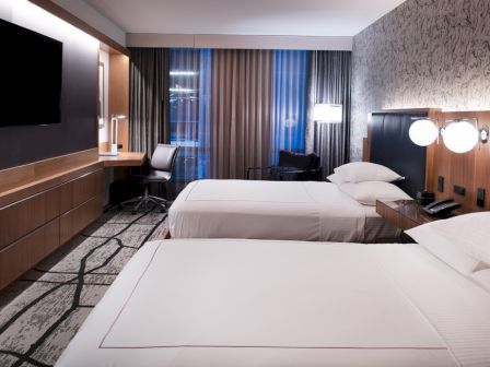 This image shows a modern hotel room with two beds, a TV, a desk and chair, and stylish decor.