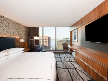 The image shows a modern hotel room with a large bed, a desk, a chair, and a TV, plus a window view of city buildings and water.