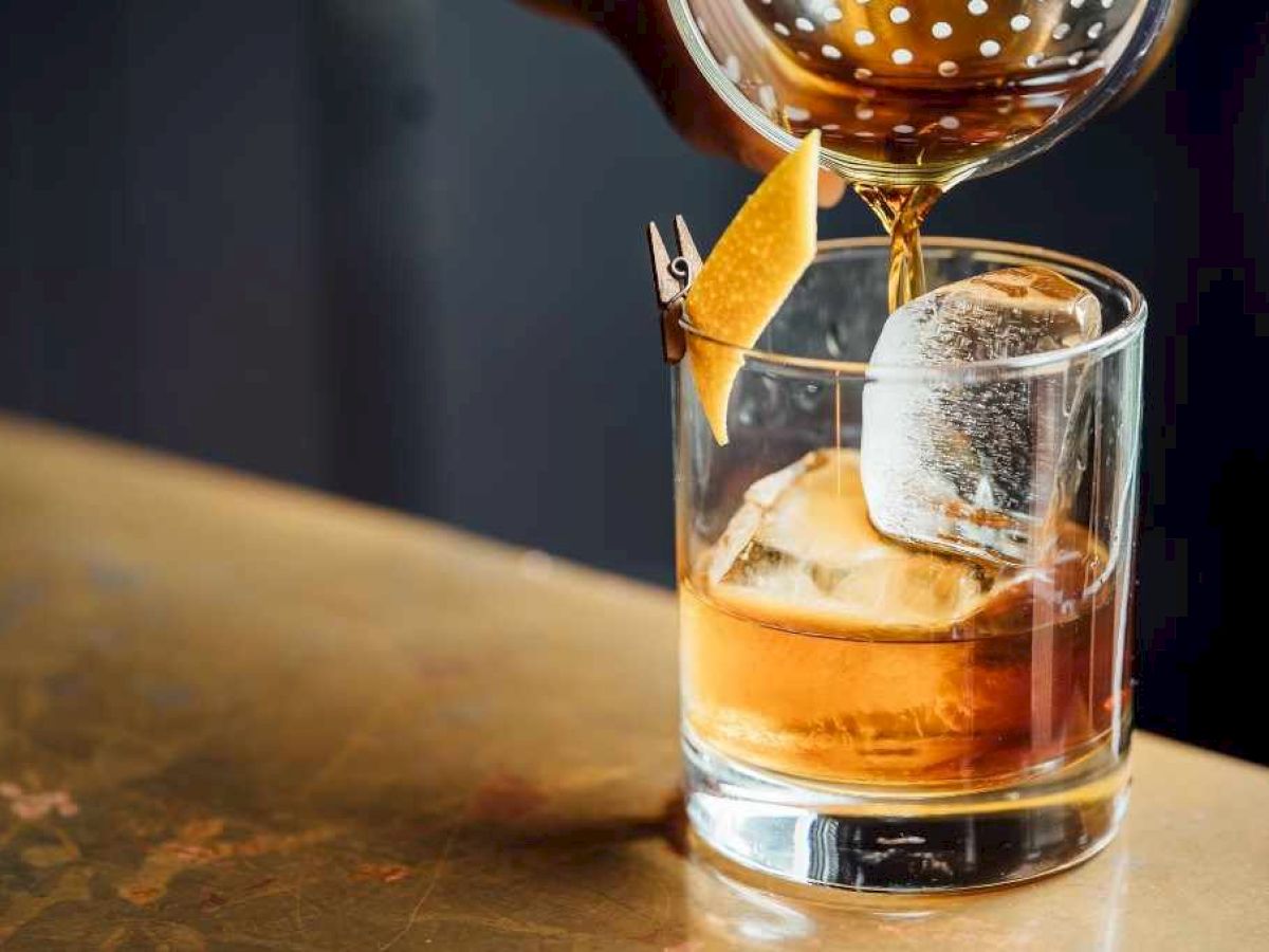 A drink is being poured into a glass with ice and garnished with a twist of citrus peel.