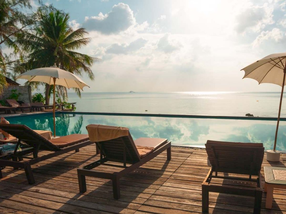 A scenic view of lounge chairs by a pool overlooking the ocean, with umbrellas and palm trees under a partly cloudy sky.