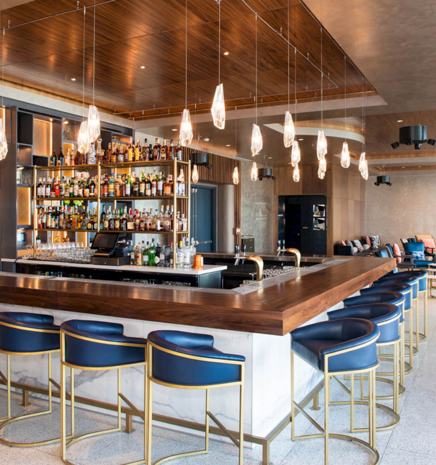 A modern bar with blue barstools, hanging lights, and a stocked back shelf, set in a spacious room with a seating area.
