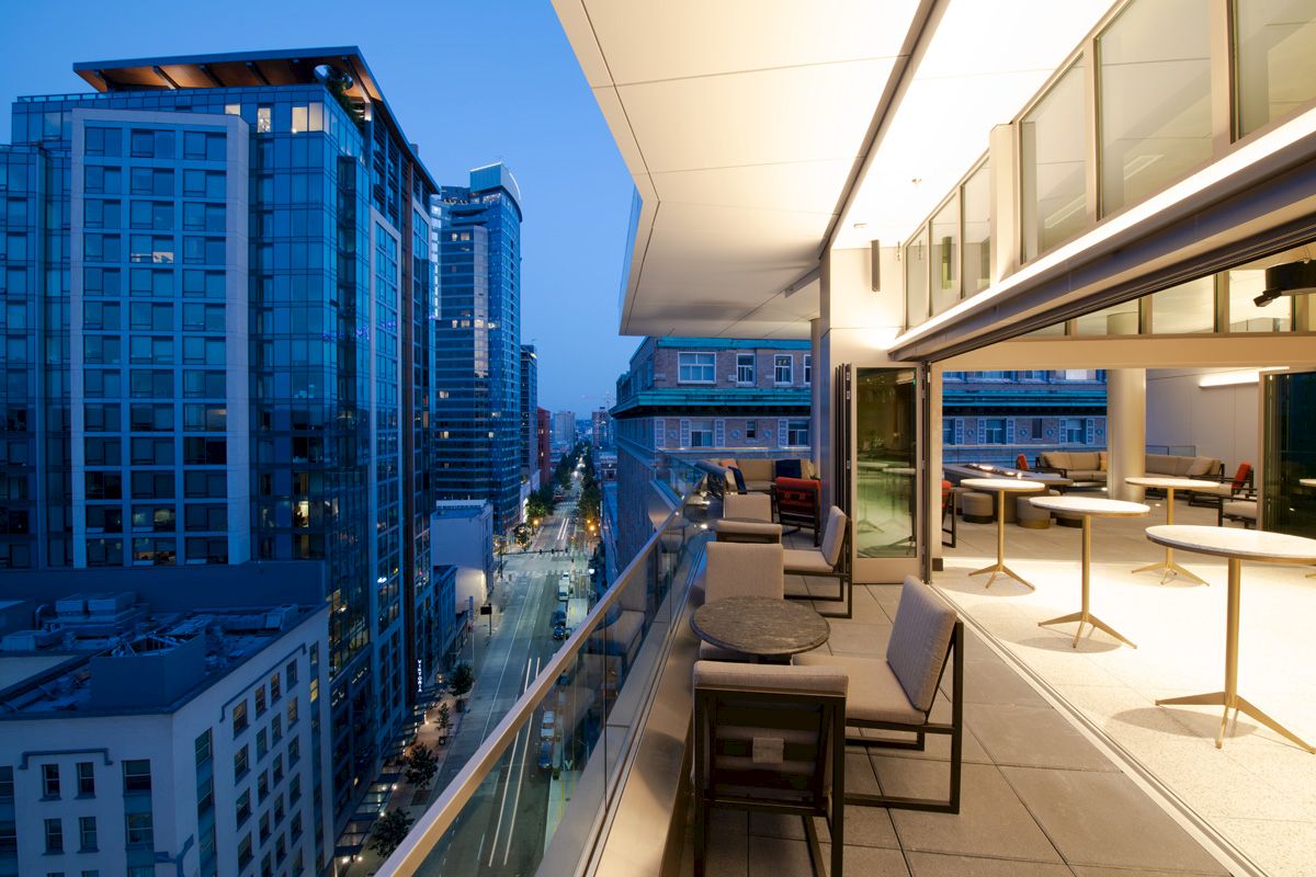 A modern rooftop terrace with outdoor seating overlooks a cityscape of tall buildings at dusk, featuring a vibrant urban environment.