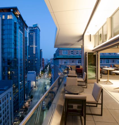 A modern balcony with seating, overlooking a cityscape with tall buildings at dusk, features large windows and a bright interior.