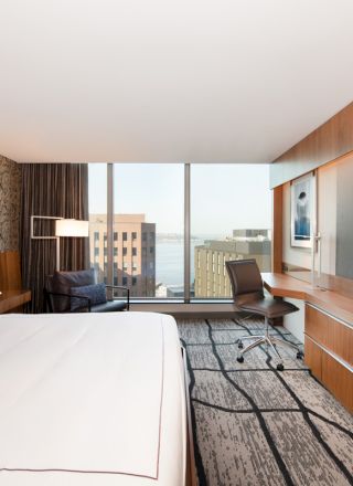 A modern hotel room with a bed, desk, and large TV, featuring a city skyline view through large windows.