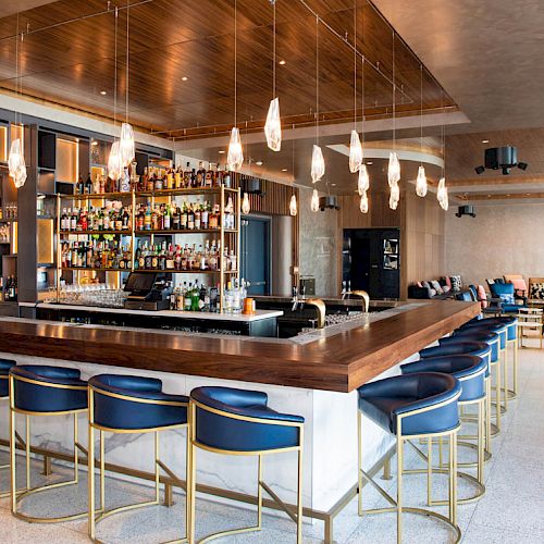 The image shows a modern bar with a central counter, blue stools, hanging lights, and a well-stocked back shelf in a stylish interior space.