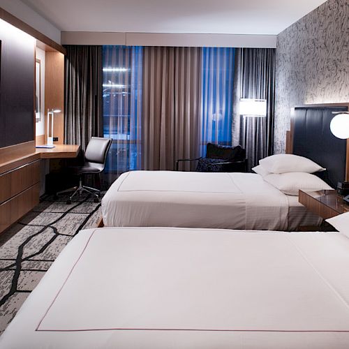 A modern hotel room with two beds, wall-mounted TV, desk, chair, lamps, and stylish decor.
