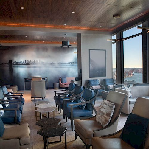 A modern lounge with comfy seating, large windows offering a scenic view, and a mural of a city skyline adorns the far wall.