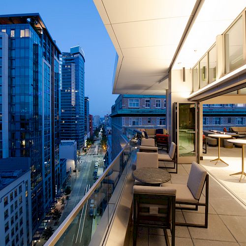 A modern balcony with seating overlooks an urban street flanked by tall buildings during twilight.