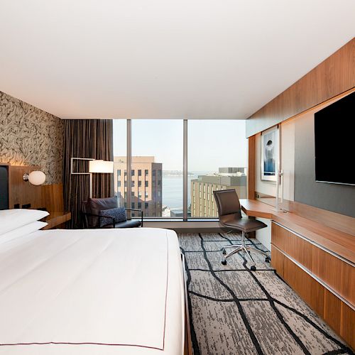 A modern hotel room with a bed, large TV, desk, and city view through a window.