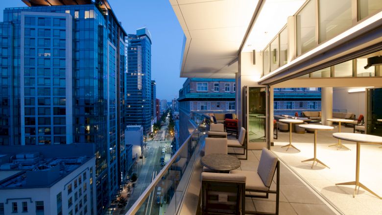 A modern cityscape features tall buildings at dusk, with a stylish rooftop terrace overlooking the street below.