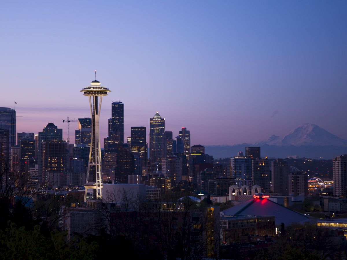 A city skyline at dusk featuring the Space Needle and buildings, with a distant view of a mountain in the background.