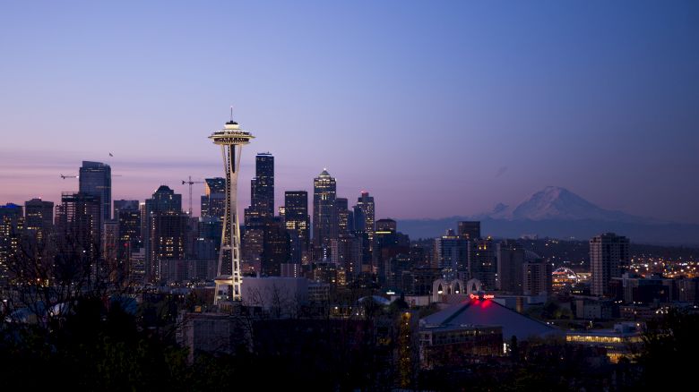 A city skyline at dusk featuring the Space Needle and buildings, with a distant view of a mountain in the background.