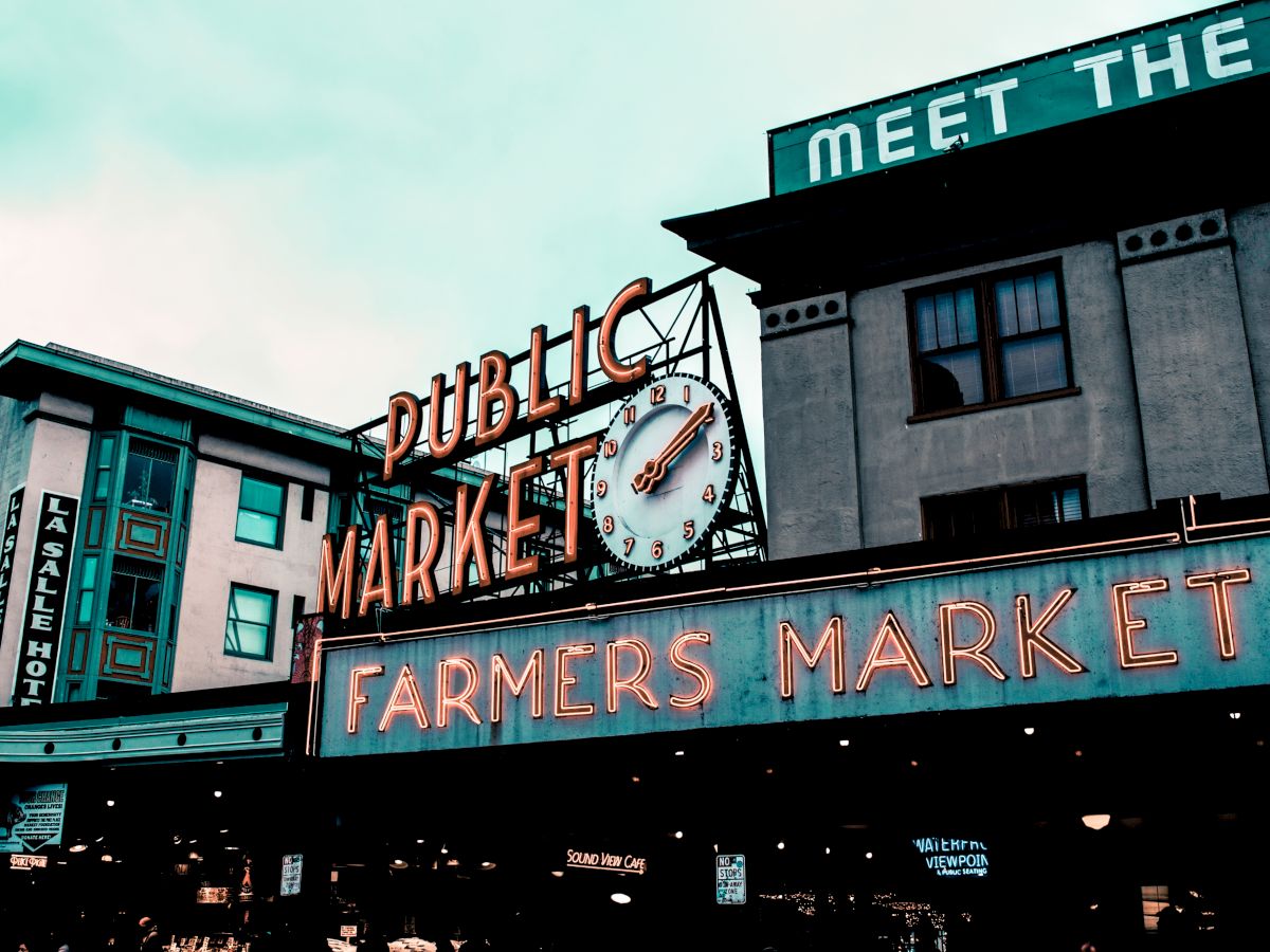 This image shows an iconic neon sign for a public market, likely a farmers market area with buildings in the background in an urban setting.