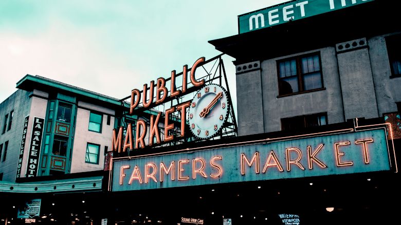This image shows an iconic neon sign for a public market, likely a farmers market area with buildings in the background in an urban setting.